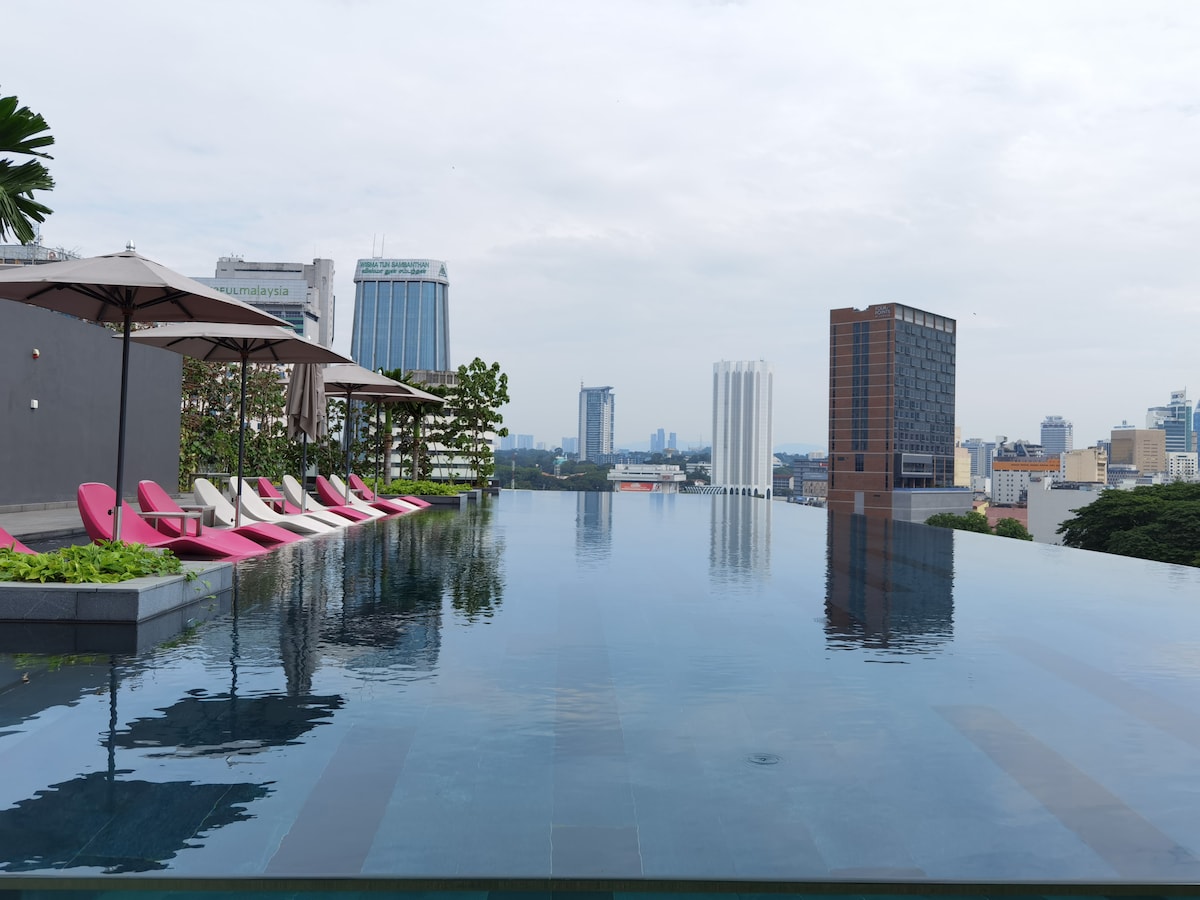 An infinity pool stretches across the rooftop, reflecting the skyline of Kuala Lumpur. White loungers with pink accents are arranged under large umbrellas, while lush greenery adds a natural touch. The tranquil water creates a serene atmosphere, inviting relaxation with city views.