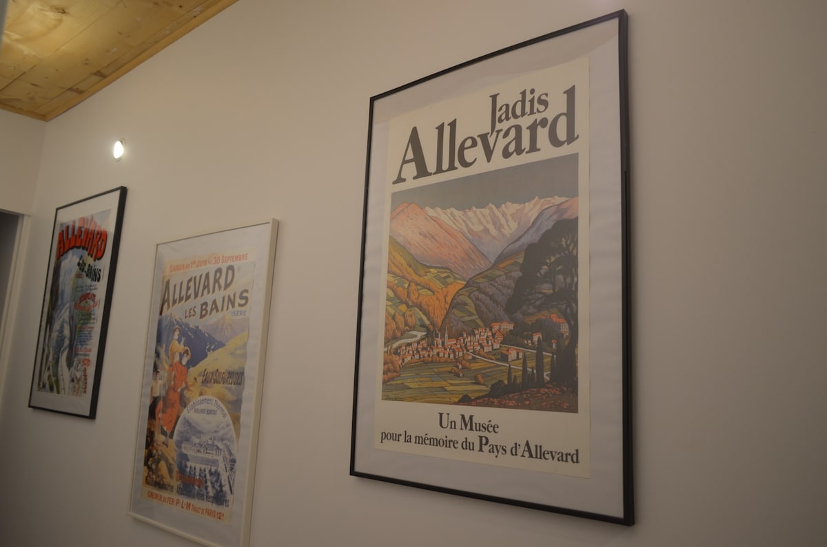 Framed vintage posters depicting Allevard are displayed on the wall, showcasing the region's scenic landscapes. The artwork features various colors and styles, contributing to the thematic decor of the space. Soft lighting highlights the details of the posters against a neutral wall.