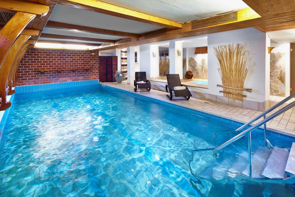 An indoor swimming pool features clear blue water, with comfortable lounge chairs positioned nearby. The ambiance is enhanced by natural wood beams and a textured stone wall. Soft lighting illuminates the area, creating a serene environment for relaxation and leisure.