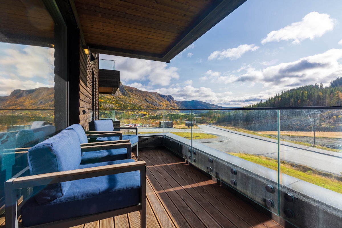A spacious outdoor terrace is furnished with comfortable blue chairs and a wooden floor. Glass railings offer unobstructed views of the surrounding mountains and landscape, inviting relaxation in a serene atmosphere.