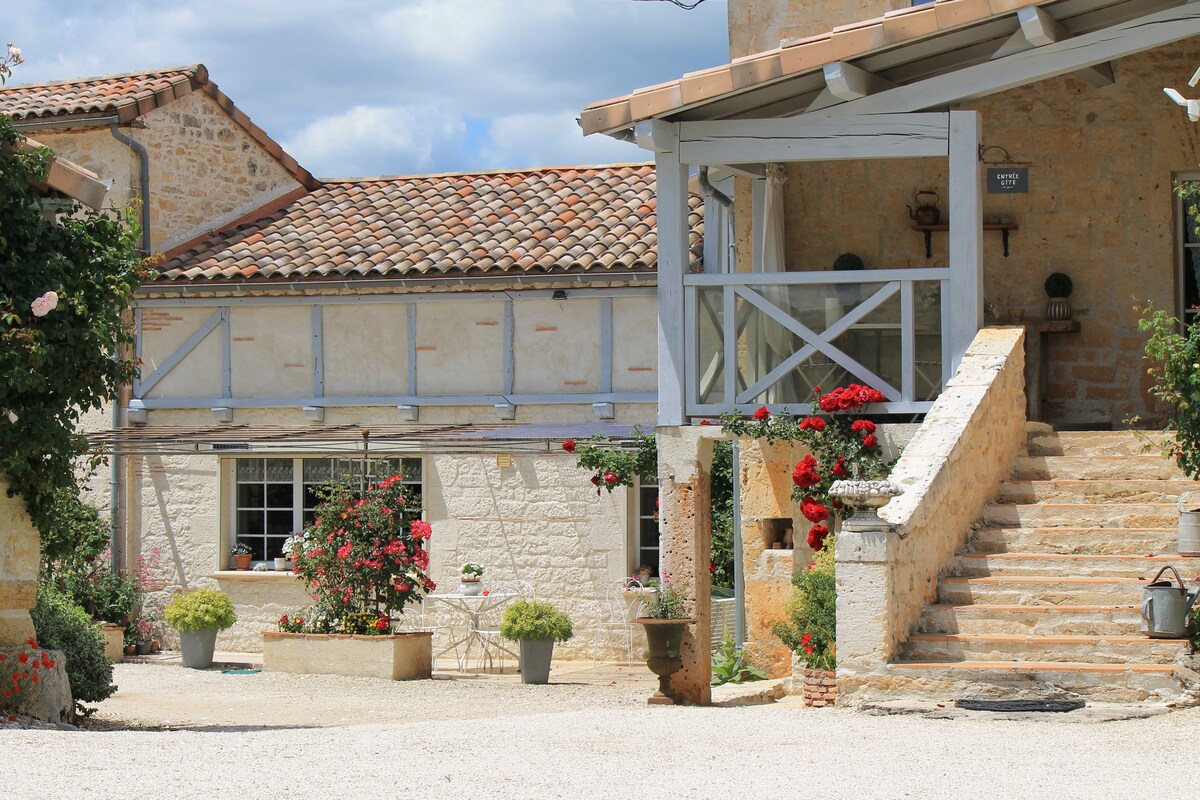 The entrance features a stone staircase leading to a charming balcony with wooden railings. Vibrant flowers line the steps and surrounding pathways, adding color to the rustic facade. A cozy seating arrangement is visible on the balcony, enhancing the inviting outdoor atmosphere.