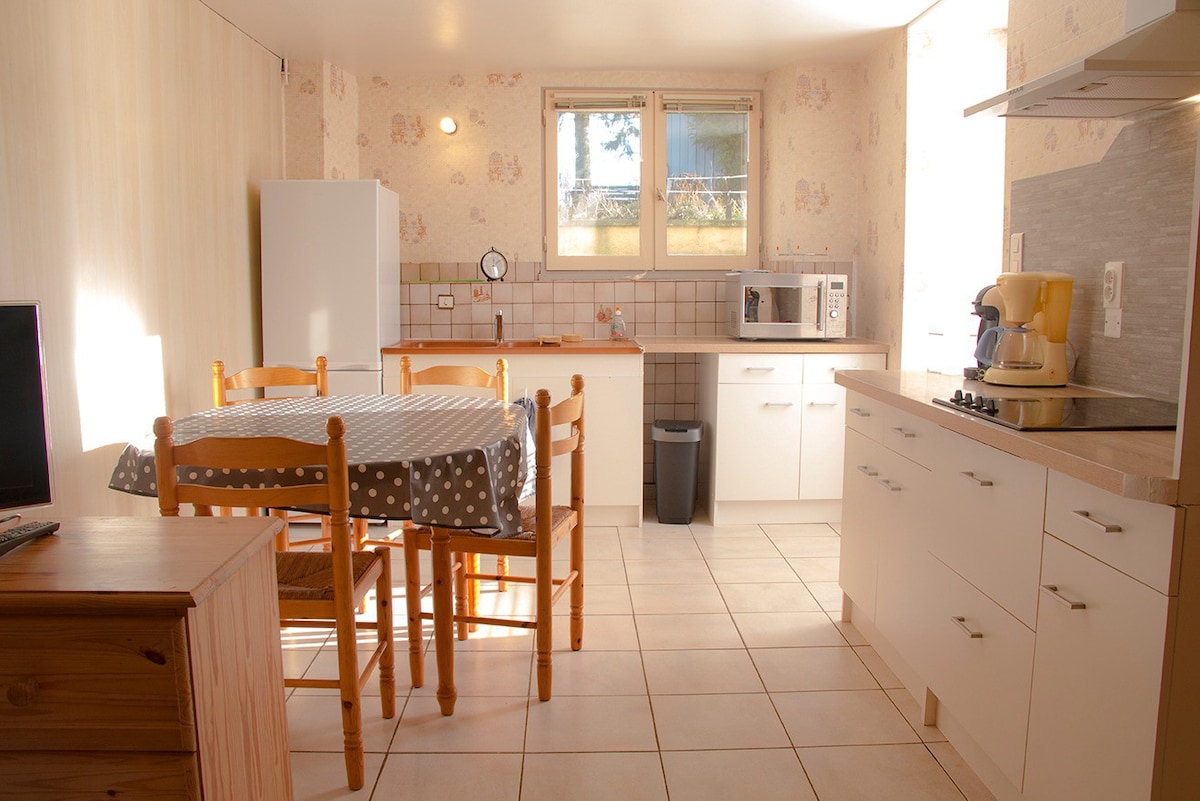 A well-equipped kitchen is shown, featuring a dining table with wooden chairs, a refrigerator, a microwave, and a countertop with modern appliances. Natural light enters through the window, illuminating the tiled floor and light wall colors.