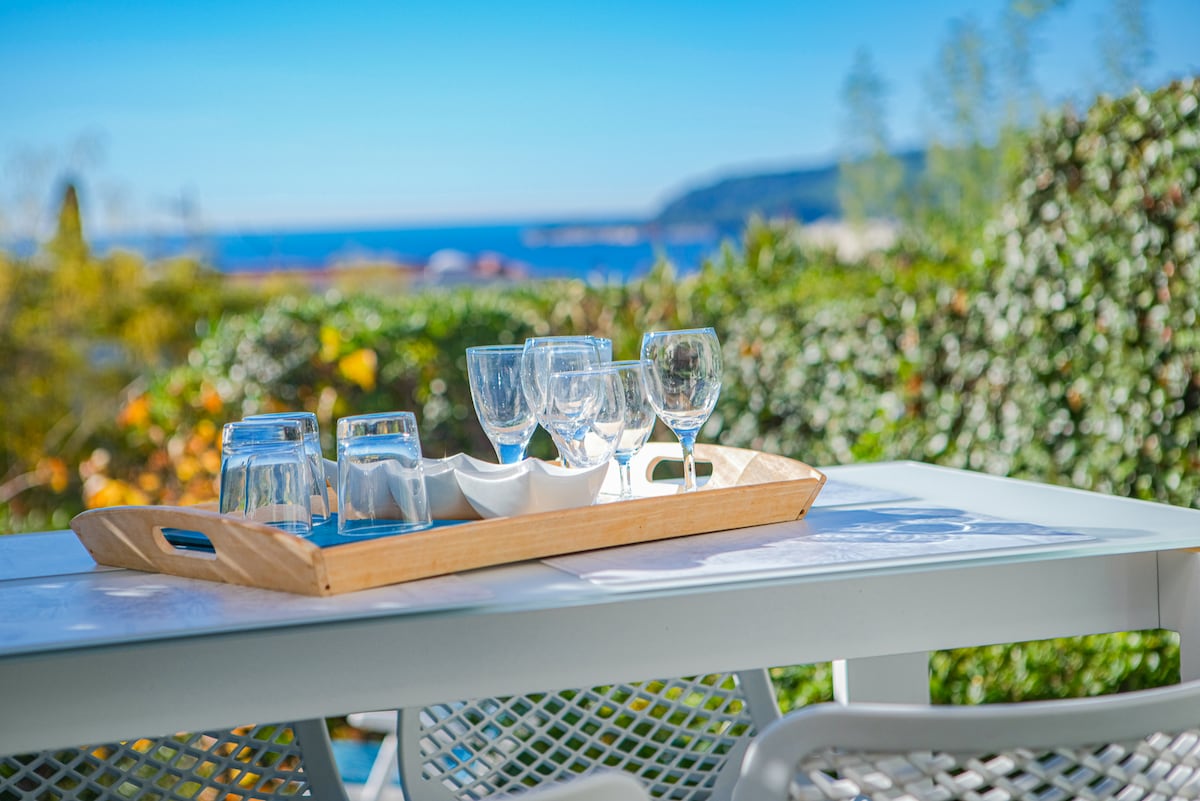 A wooden tray displays glasses and cup sets arranged on a table, set against a backdrop of lush greenery and a view of the sea. Natural light enhances the tranquil setting, inviting relaxation.