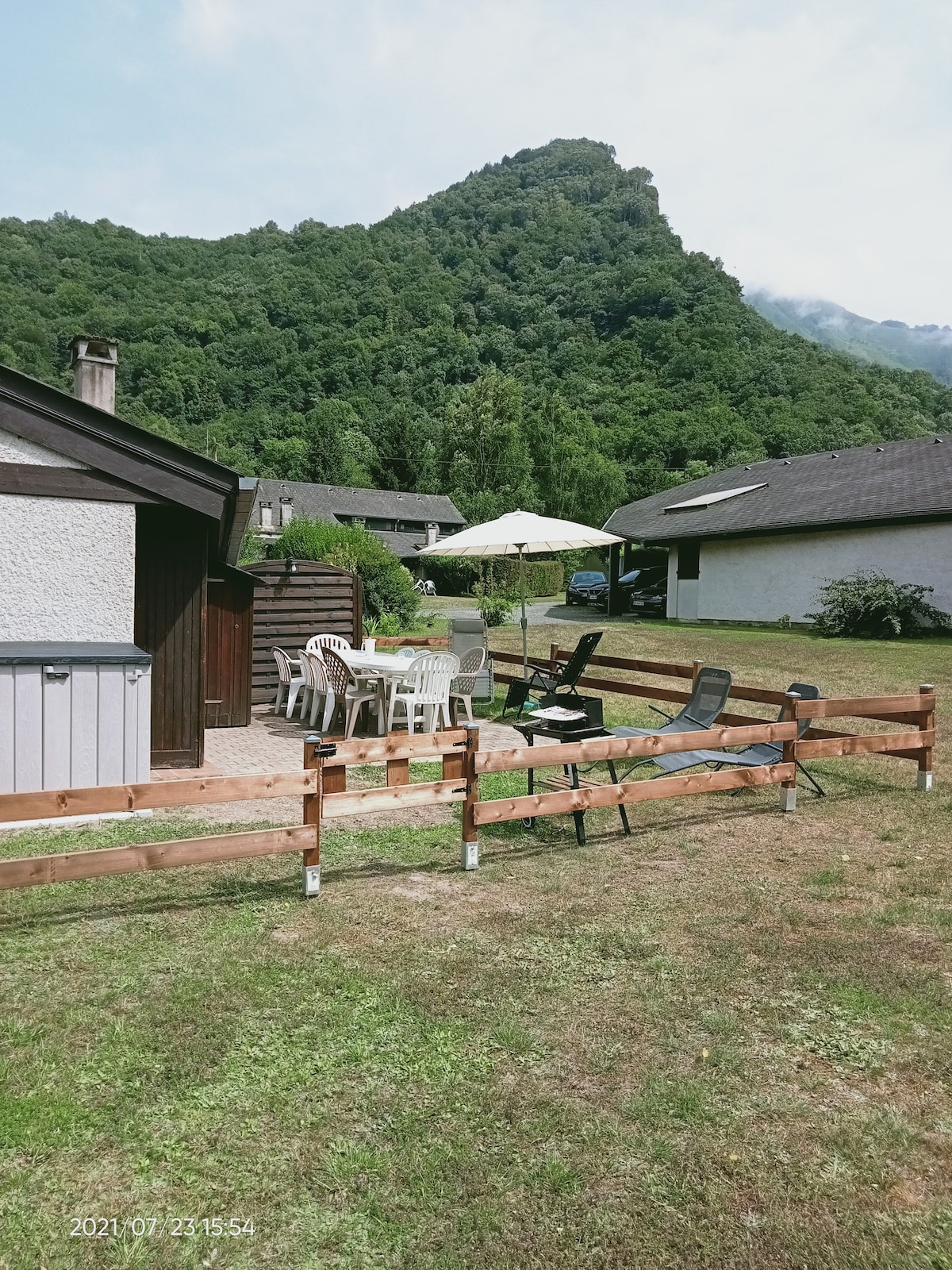 An outdoor terrace is seen with a dining table, white chairs, and a sun umbrella, creating a space for relaxation. Surrounding greenery includes a hillside in the background, providing a natural setting. Fencing adds a defined border to the grassy area nearby.