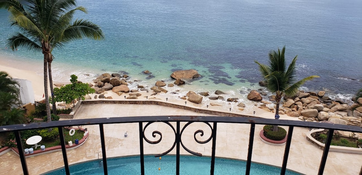 A view from a private balcony showcases a serene beach with gentle waves lapping at the shore. Palm trees frame the scene, and smooth stones are visible in the clear blue waters, creating a peaceful and relaxing coastal atmosphere.