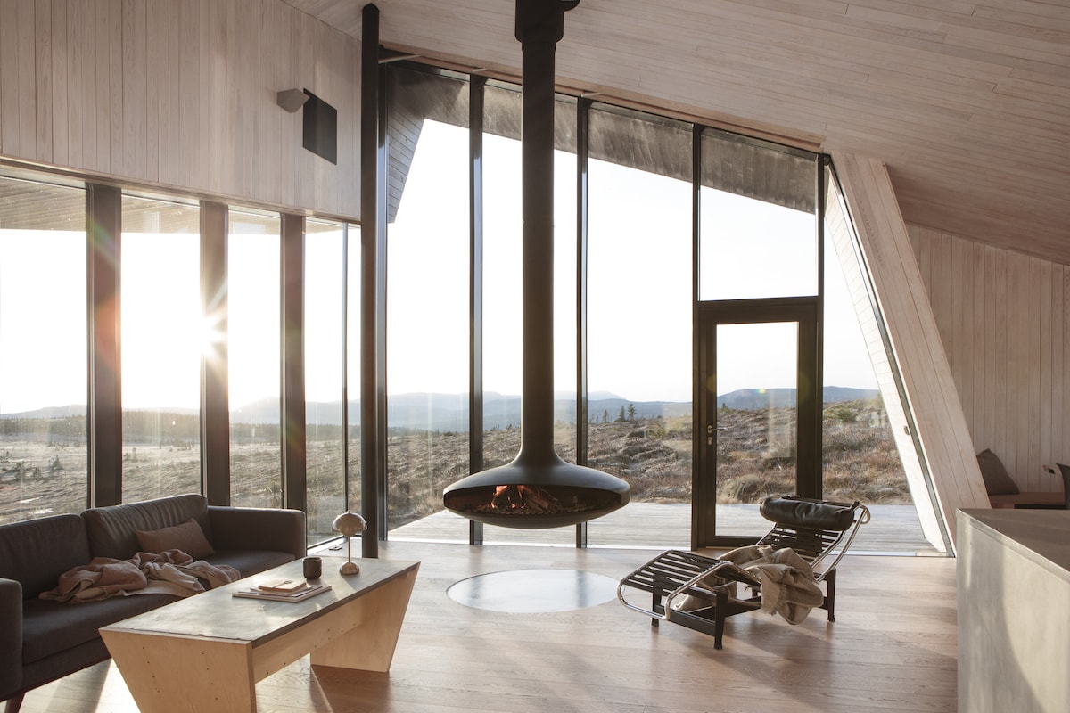 The spacious living room features large floor-to-ceiling windows, offering expansive views of the surrounding nature. A modern hanging fireplace is centered in the room, with a lounge chair positioned nearby. A simple wooden table holds decorative items, enhancing the contemporary design of the space.