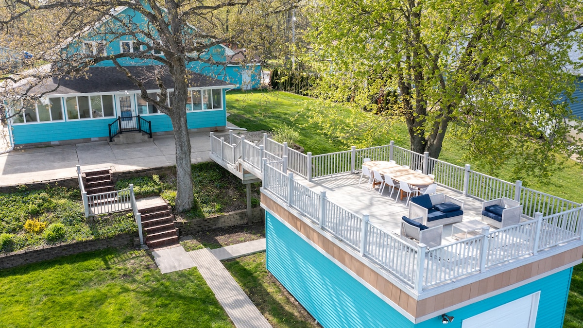 A spacious rooftop deck is featured atop a vibrant blue house, equipped with outdoor seating. Nearby, a well-maintained patio area and grassy lawn are surrounded by mature trees, providing a tranquil outdoor space for relaxation and gatherings.