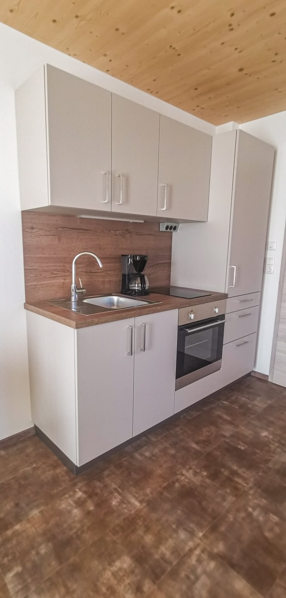 A compact kitchen area features modern cabinetry in neutral tones. A stainless steel sink, an oven, and a stovetop are included, along with a coffee maker on the counter. The wood paneling above adds warmth to the functional space.