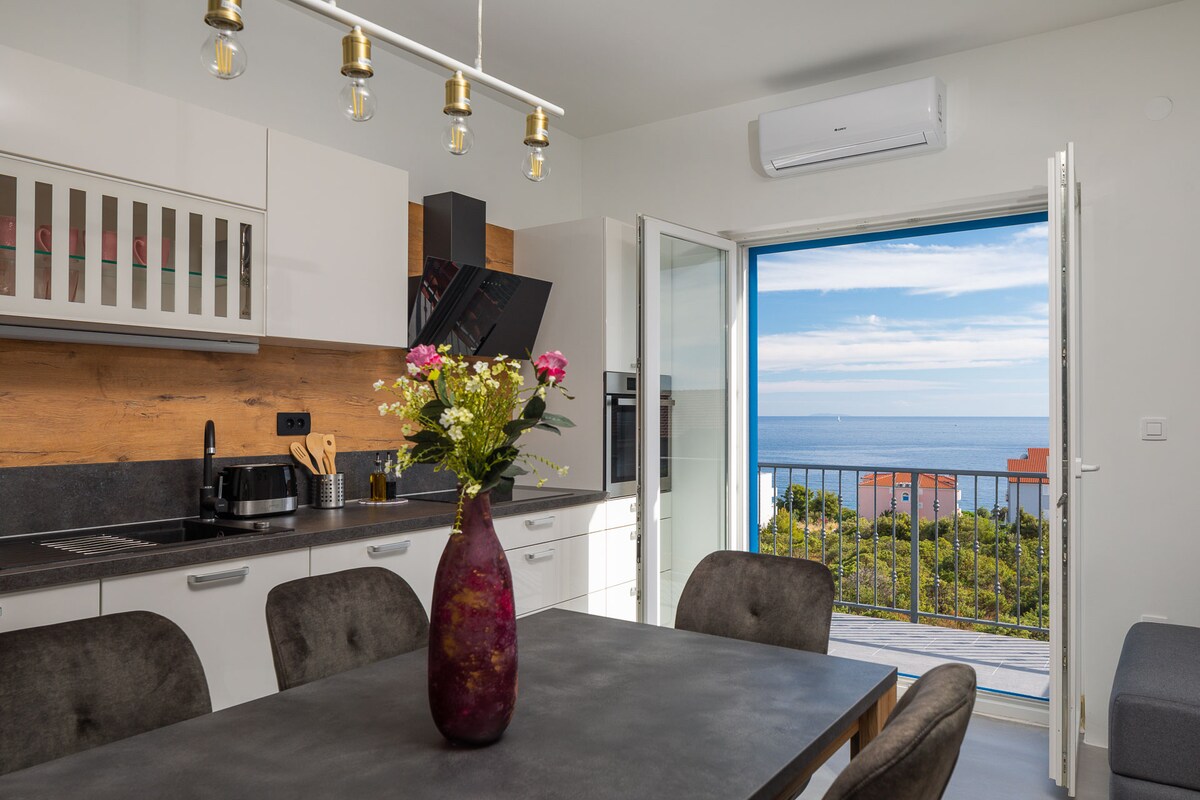 A modern kitchen is featured with sleek cabinetry and a dining table, adorned with a vase of flowers. Large glass doors open to a balcony, revealing a panoramic sea view and bright skies. Air conditioning ensures comfort within the space.