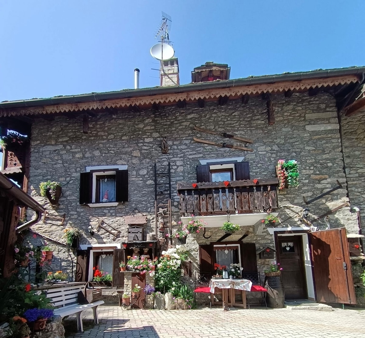 The exterior of a charming stone building is displayed, featuring dark wooden shutters and decorative flower boxes filled with vibrant blooms. A balcony adorned with plants overlooks a quaint courtyard, while rustic wooden benches invite relaxation in the sunlight.