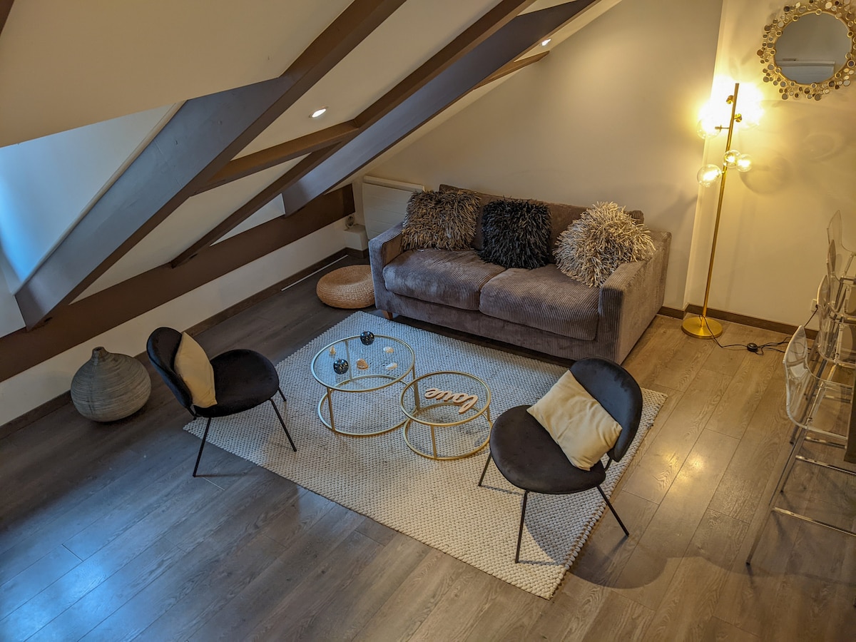 A cozy seating area is presented with a plush sofa adorned with decorative cushions. Two sleek chairs frame a glass coffee table, all positioned on a textured area rug. Soft lighting is provided by a modern floor lamp, accentuating the sloped ceiling and wooden beams.