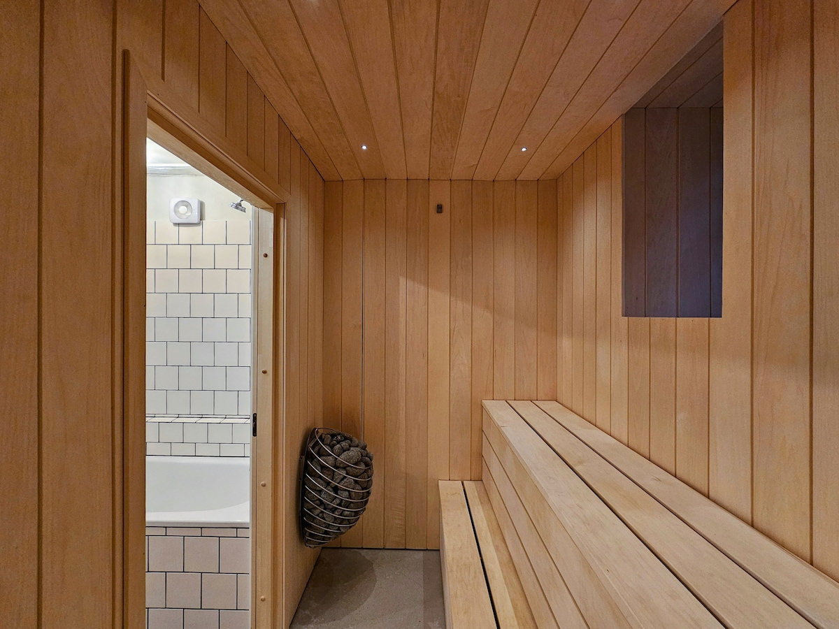 A wooden sauna interior features benches along the walls, offering a serene space for relaxation. A small window allows soft light to enter, while a basket sits neatly in the corner. The adjoining bathroom is visible through an open doorway.