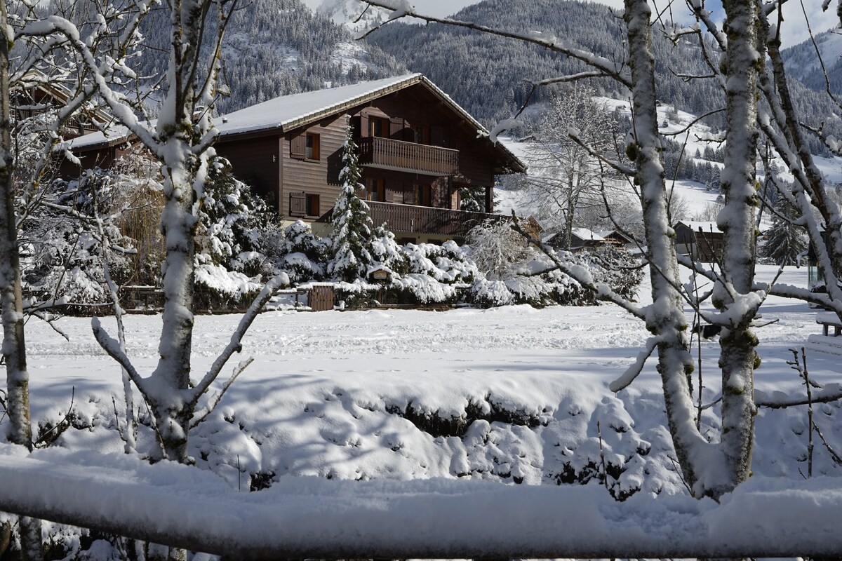 A wooden chalet is surrounded by a serene winter landscape, with snow covering the ground and trees. The chalet features multiple balconies and is nestled among evergreen trees, with a backdrop of snow-capped mountains visible in the distance.