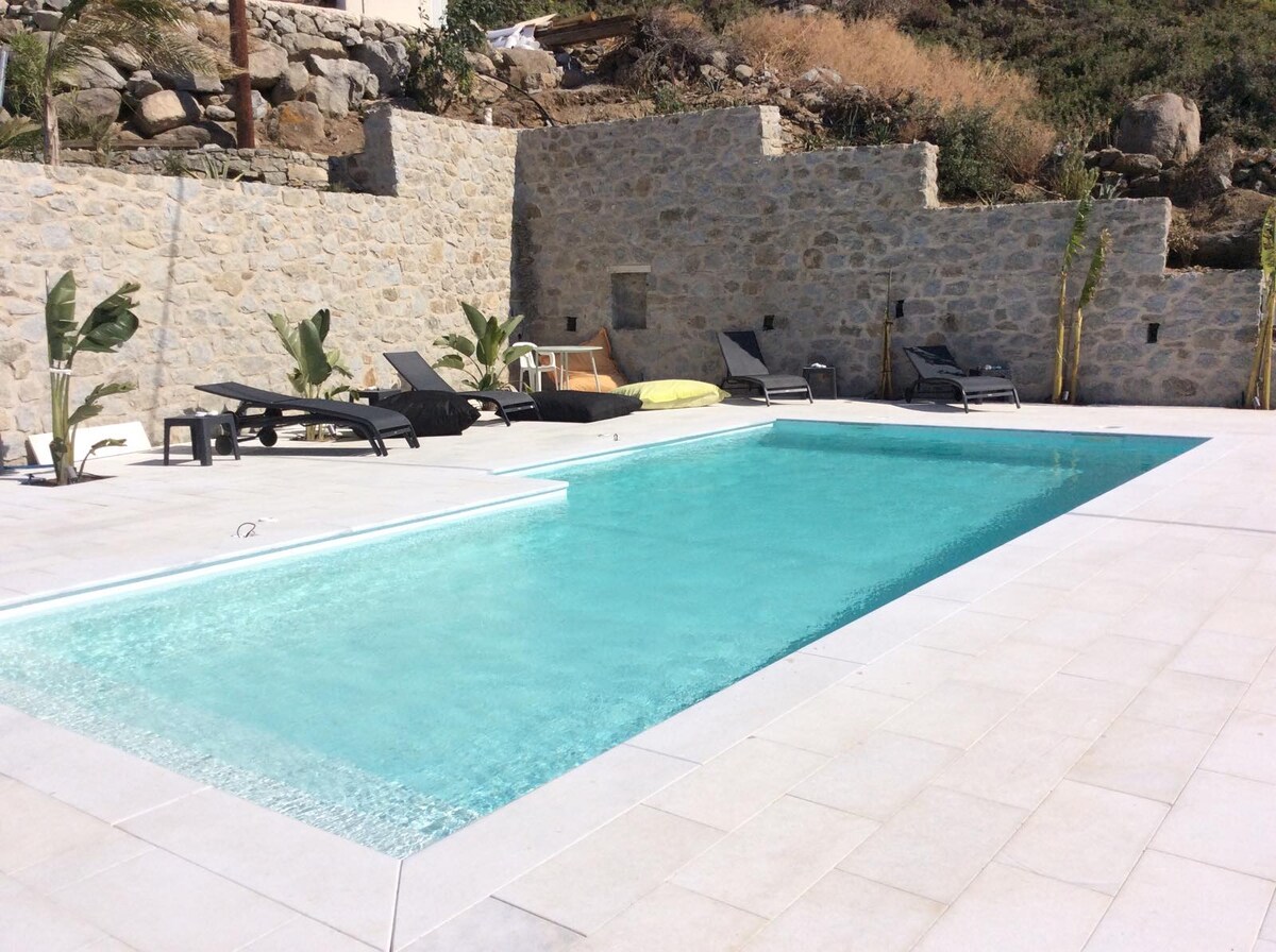 A serene pool area features a turquoise swimming pool surrounded by stone walls and paved flooring. Several lounge chairs are positioned nearby, with tropical plants enhancing the space's natural ambiance.