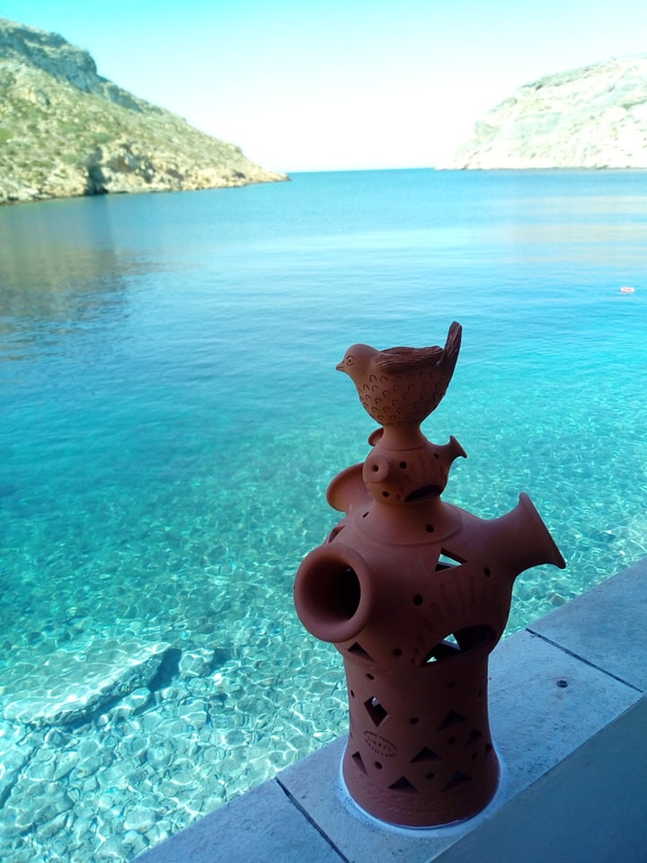 A decorative terracotta sculpture featuring birds and fish is positioned on a ledge overlooking a clear blue sea. The water reflects shades of turquoise, and rocky hills are visible in the background, enhancing the serene coastal view.