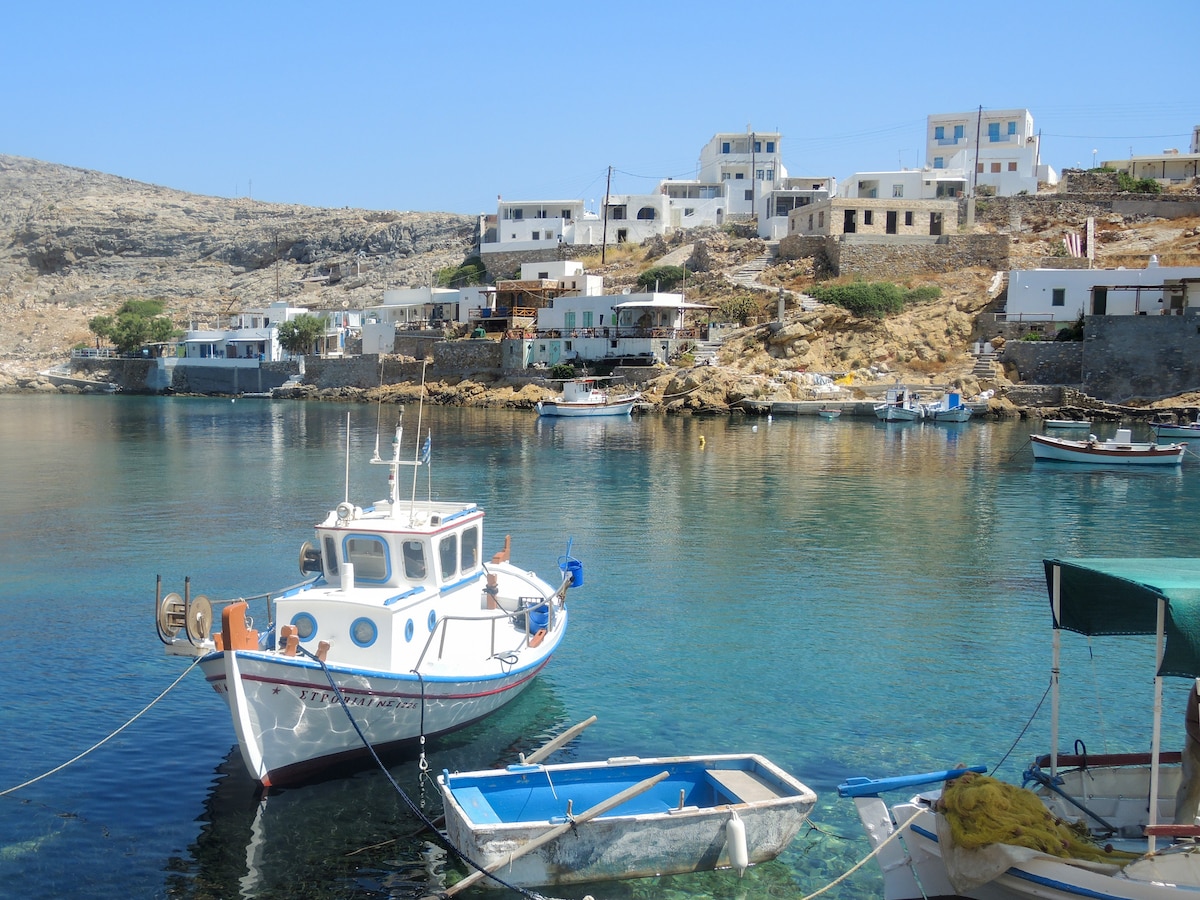 A tranquil scene of a fishing village by the sea is depicted, featuring moored boats on calm waters. Whitewashed buildings rise on the hillside, with rocky terrain in the background, and clear blue skies above. The area conveys a serene coastal atmosphere.