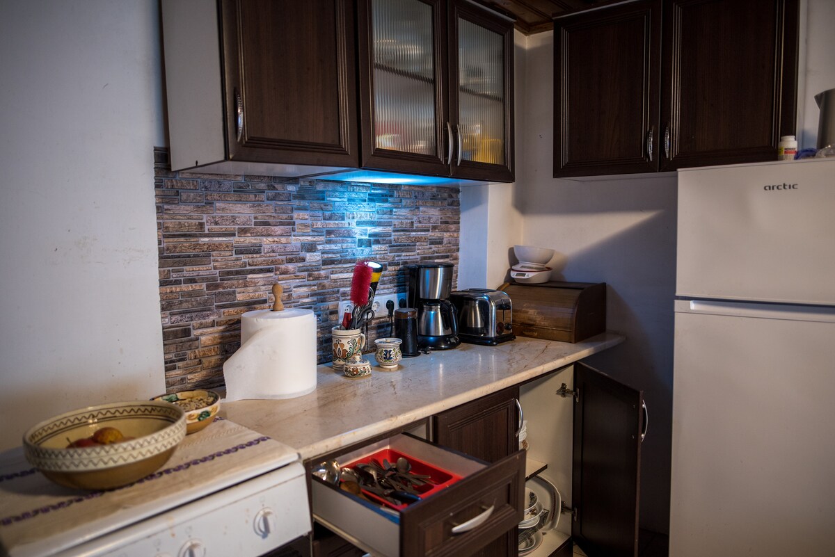 The kitchen area features wooden cabinetry and a countertop with modern appliances, including a coffee maker and kettle. A neatly organized drawer is visible, along with a full-size refrigerator. Subtle lighting highlights the textured stone backsplash, creating a functional cooking space.