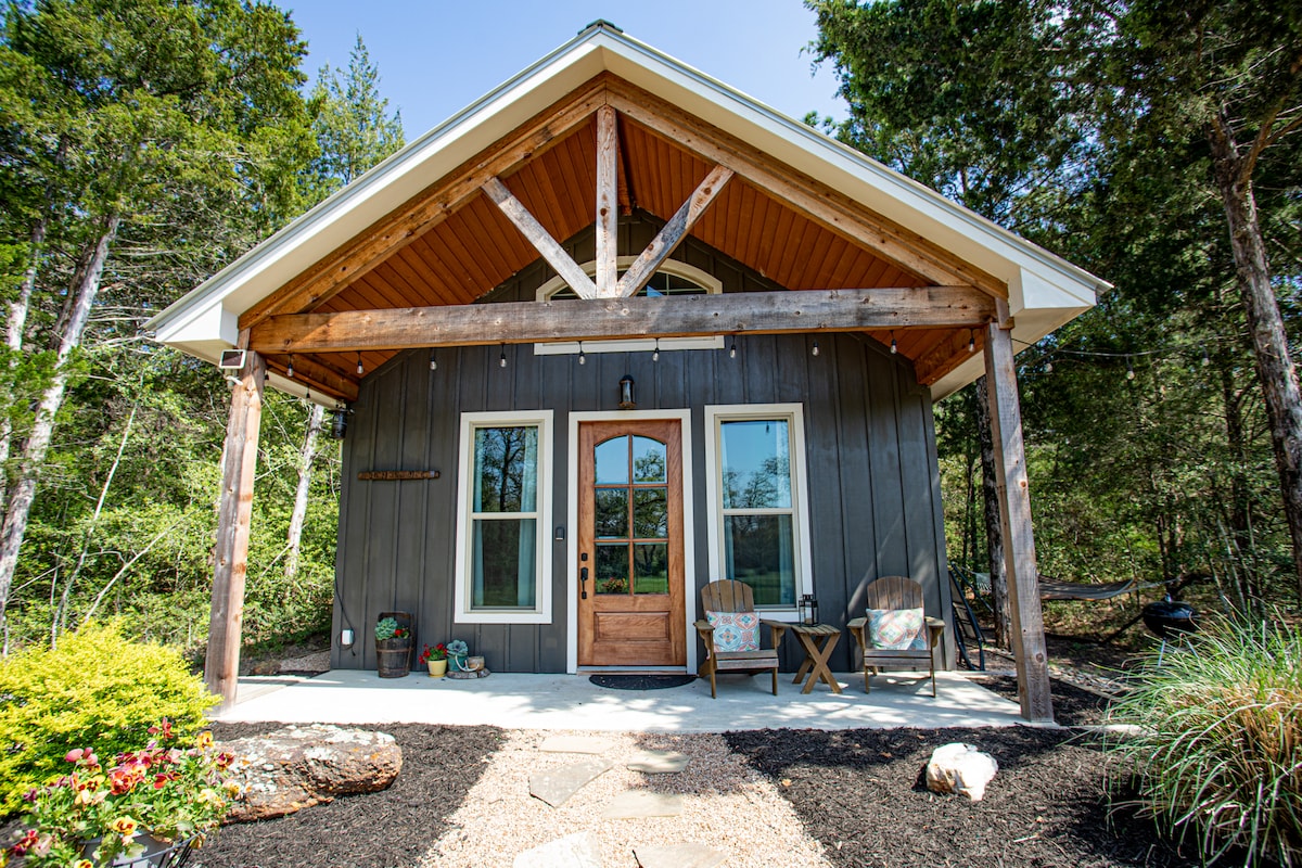 The cottage exterior features a wooden door at the center, framed by two large windows. A covered porch is highlighted by two chairs, inviting relaxation. Natural landscaping with stones and colorful flowers surrounds the entrance, set against a backdrop of trees under a clear blue sky.