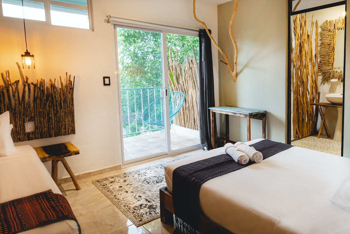 A bright room features a queen-size bed with white linens, complemented by a single bed. Natural light enters through large sliding doors that lead to a private balcony. A rustic wooden accent wall adds character, while the floors are tiled for easy cleaning.
