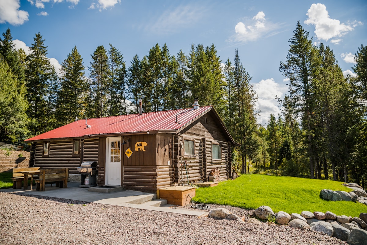 A vintage log cabin is surrounded by tall pine trees, showcasing a red metal roof and wooden exterior. A gravel pathway leads to the entrance, and an outdoor grill is positioned near a seating area. Lush green grass enhances the outdoor space.