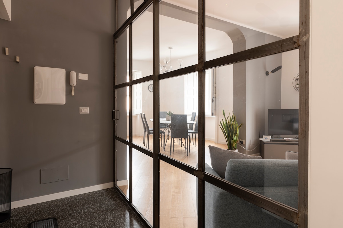 The image shows a modern interior space with a glass door providing a view into a well-lit living area. A cozy sofa is visible in the foreground, while a dining table with chairs is seen in the background, surrounded by bright natural light from the windows.