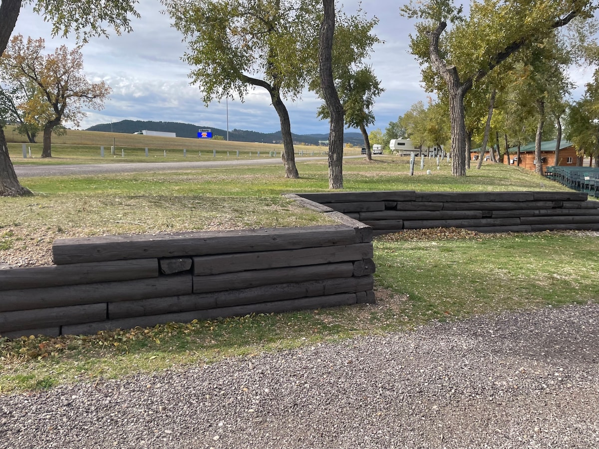 A landscaped area features wooden retaining walls made of dark timber, defining a grassy space. Nearby trees provide shade, and a gravel path leads through the area, offering easy access. The backdrop includes rolling hills and open fields, showcasing a serene outdoor setting.