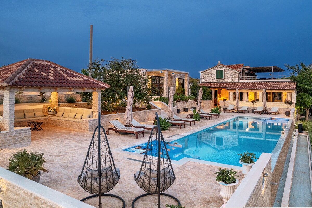 An outdoor space features a spacious swimming pool surrounded by lounge chairs. A covered seating area with a subtle tile pattern offers comfortable seating. Lush greenery and well-maintained landscaping enhance the serene atmosphere, while a rustic stone building adds character to the surroundings.