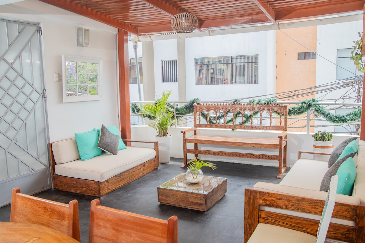 A covered terrace area features wooden seating arranged around a low coffee table. Soft cushions in shades of teal and gray add comfort, while potted plants bring a touch of greenery. The space is well-lit and offers a view of the surrounding buildings.