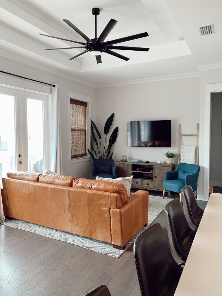 A cozy living area features a leather couch facing a wall-mounted TV. Two accent chairs are situated to the side, and a ceiling fan hangs above. Natural light enters through glass doors, with woven shades providing subtle texture. A decorative plant adds a touch of greenery.