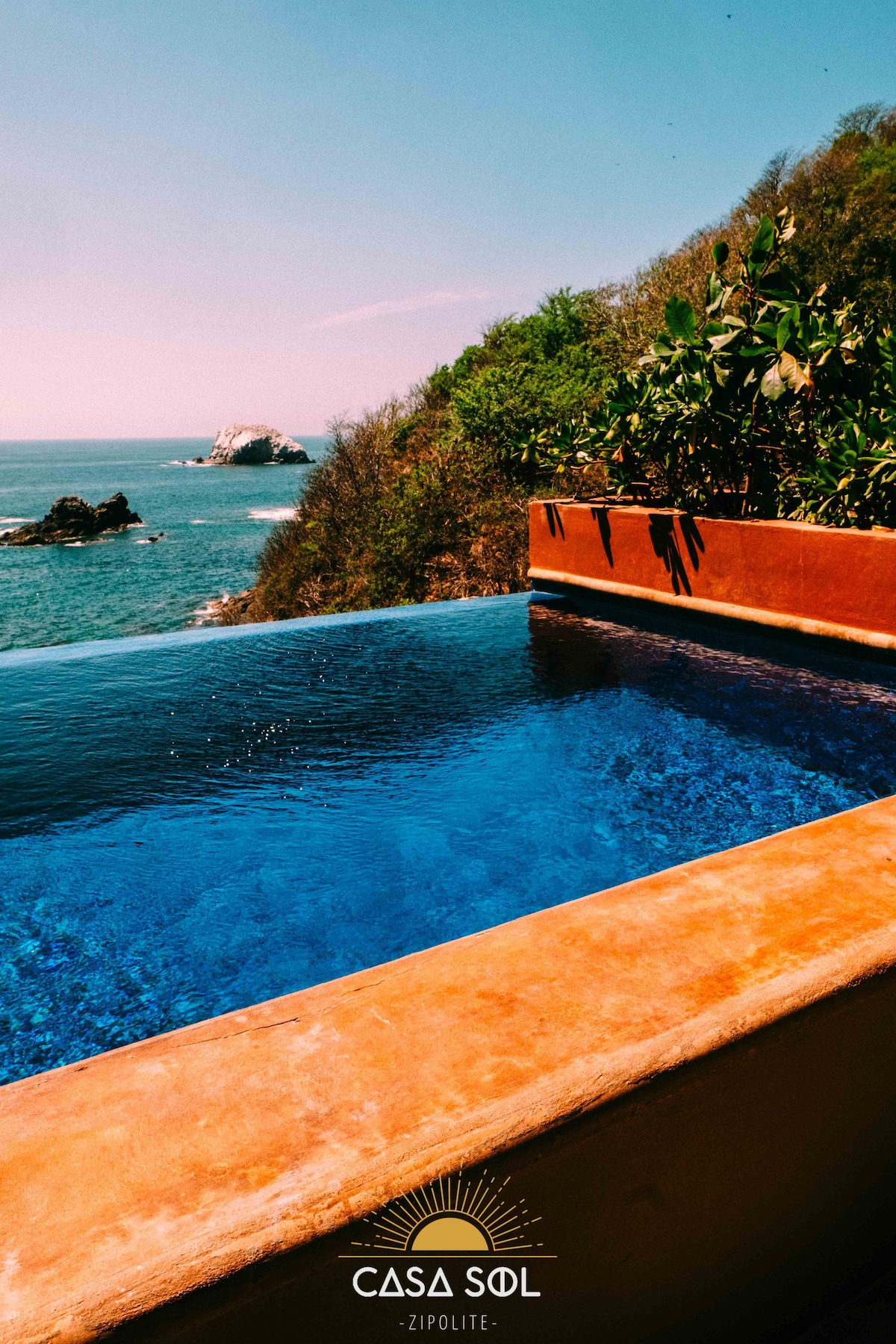 An infinity pool is situated at the edge of a cliff, offering unobstructed views of the Pacific Ocean and nearby rock formations. Sunlight glimmers on the water's surface, surrounded by lush greenery and natural textures of the surrounding landscape.