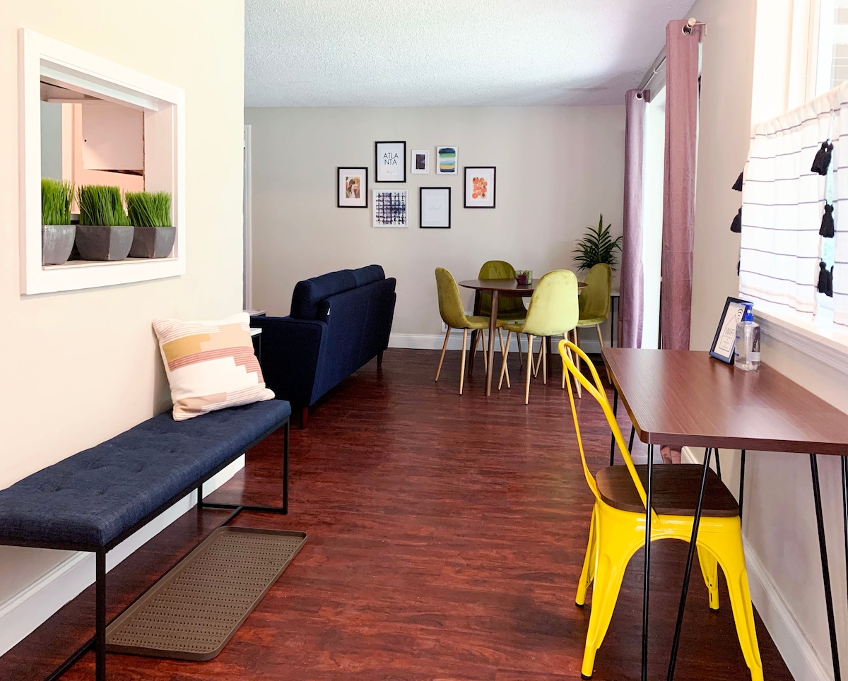 A spacious living area features a comfortable navy sofa and a dining table surrounded by colorful chairs. A bench with decorative cushions adds a cozy touch. Natural light streams through the windows, illuminating the modern decor and artwork on the walls.