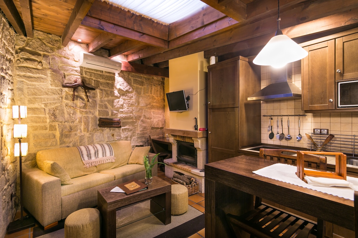 A rustic living area features exposed stone walls and wooden beams. A comfortable sofa is positioned next to a modern coffee table. A flat-screen TV is mounted above a cozy fireplace, while a well-equipped kitchen area showcases wooden cabinetry and essential cooking utensils.