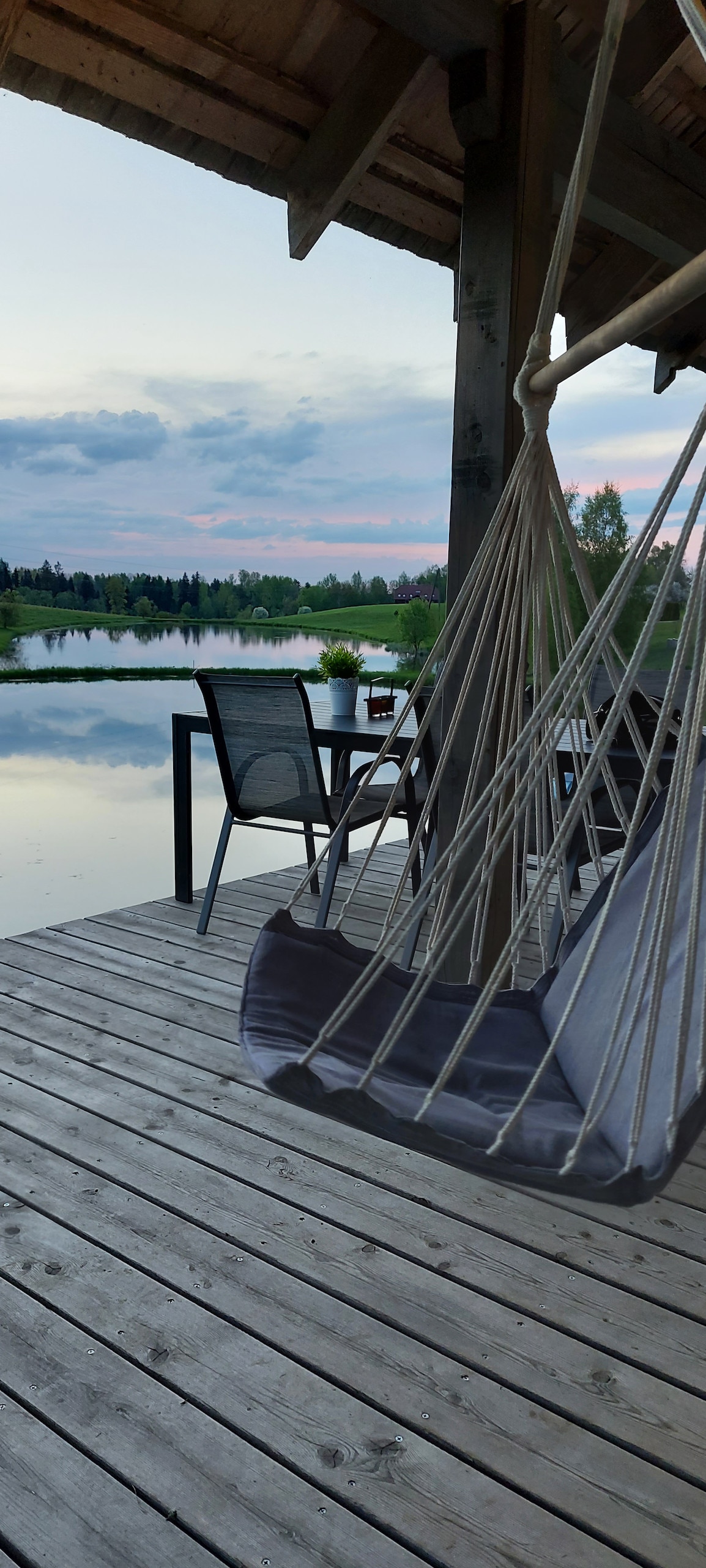 A wooden deck overlooking a serene body of water is equipped with a hammock and a chair. Soft colors of the evening sky are reflected in the calm water, while lush greenery surrounds the scene, providing a peaceful outdoor setting.