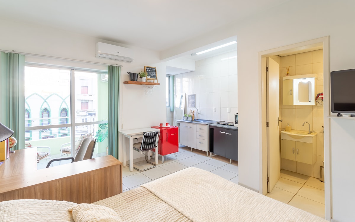 A well-lit studio apartment features a spacious sleeping area with light-colored bedding. A small dining table with red chairs is situated near the window, while a kitchen area with modern appliances is visible. A separate bathroom is adjacent, providing convenience.
