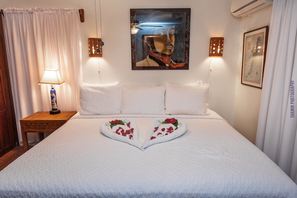 A spacious bed is centered in the room, adorned with white linens and decorative pillows. Two heart-shaped towel arrangements made with fresh flowers rest on the bed. Soft lighting is provided by bedside lamps, while a mirror and artwork enhance the cozy atmosphere.
