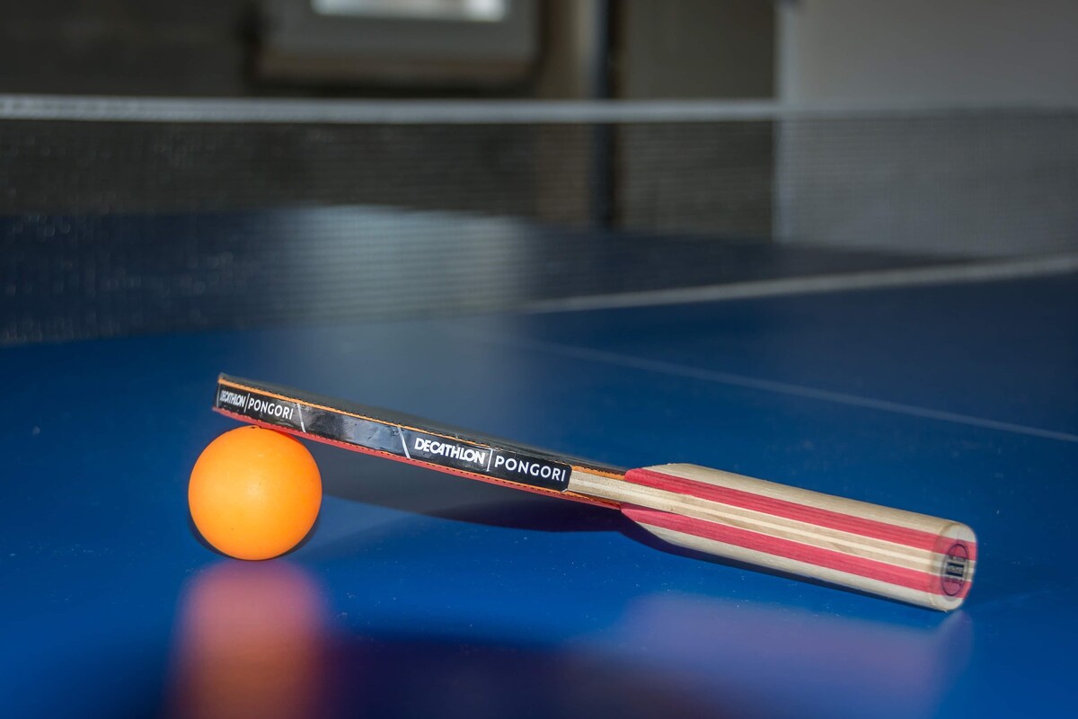 A table tennis paddle and a bright orange ball are positioned on a blue table. The paddle features a red and black design, resting close to the ball, ready for play. The table's smooth surface is highlighted, with a net visible in the background.