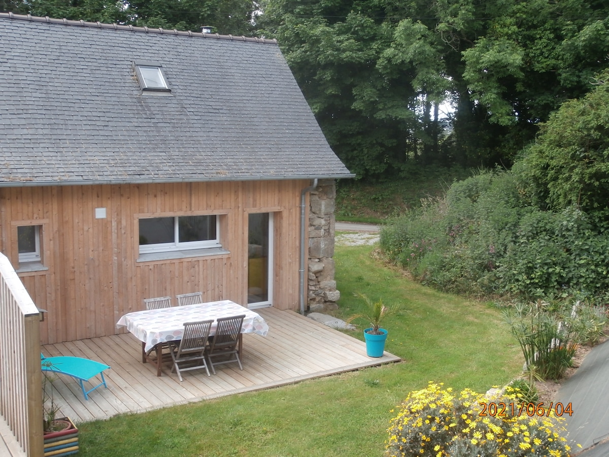 A charming wooden cottage features a spacious deck with a dining table and chairs. A colorful potted plant adds a touch of brightness. Lush greenery surrounds the area, enhancing the natural setting while a pathway leads further into the landscape.