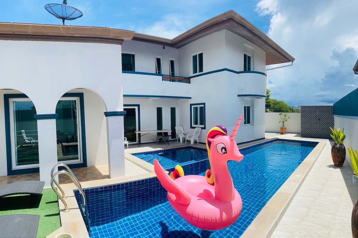 A spacious outdoor area features a swimming pool with a shiny blue surface. A large pink inflatable float in the shape of a unicorn adds a playful touch. The villa's white exterior, accented with blue lines, provides a welcoming backdrop with various seating options around the pool.