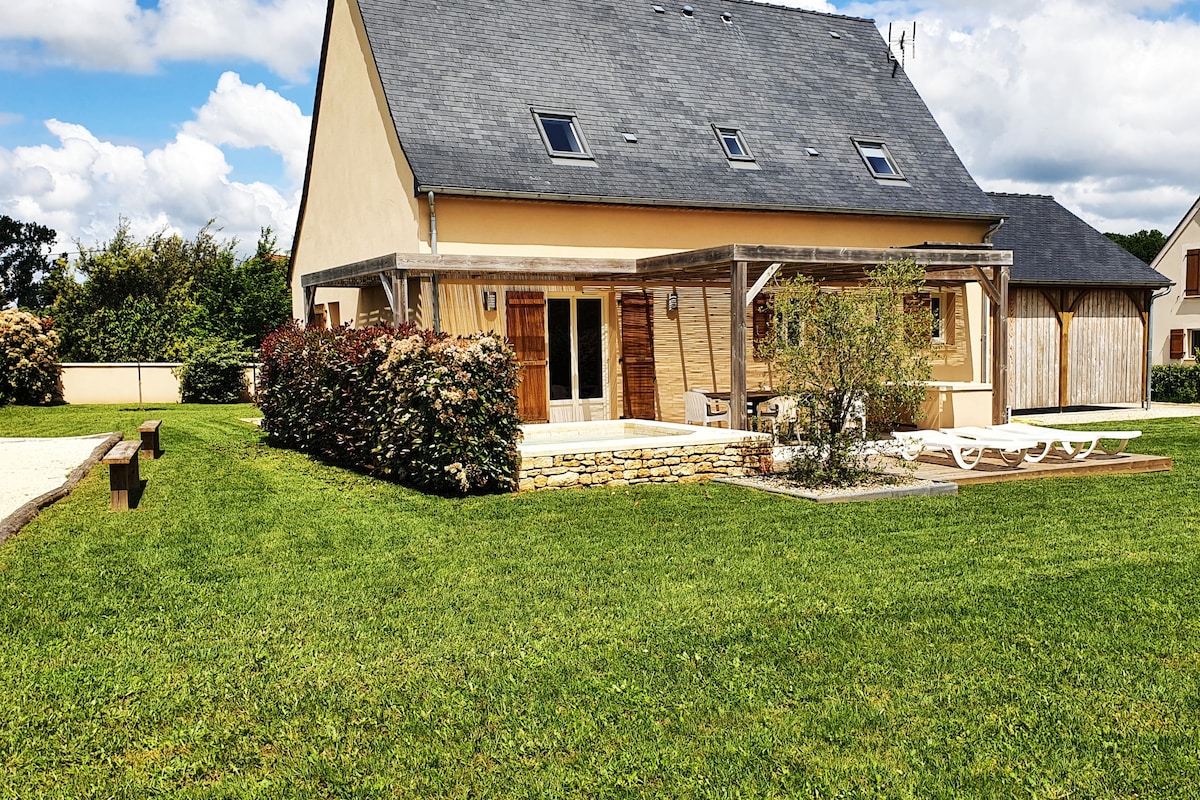 The exterior of the property is shown, featuring a two-story house with a grey roof and light yellow walls. A covered terrace is visible, along with a small pool area. Well-maintained grass surrounds the house, and landscaped hedges provide a natural boundary.