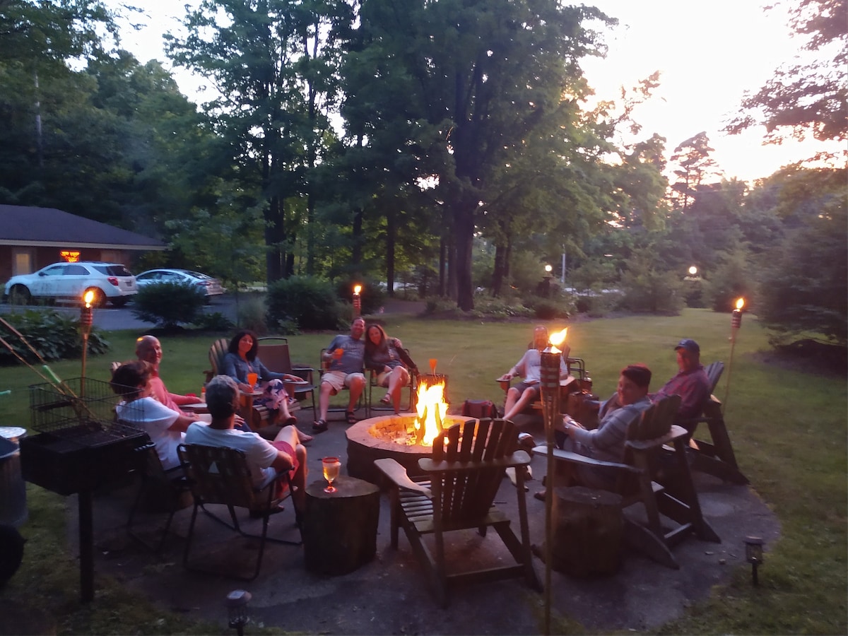 An outdoor gathering features several individuals seated around a fire pit, surrounded by tall trees. Adirondack chairs are arranged in a circle, with tiki torches providing a soft glow as dusk approaches. The tranquil setting emphasizes the inviting atmosphere of camaraderie.