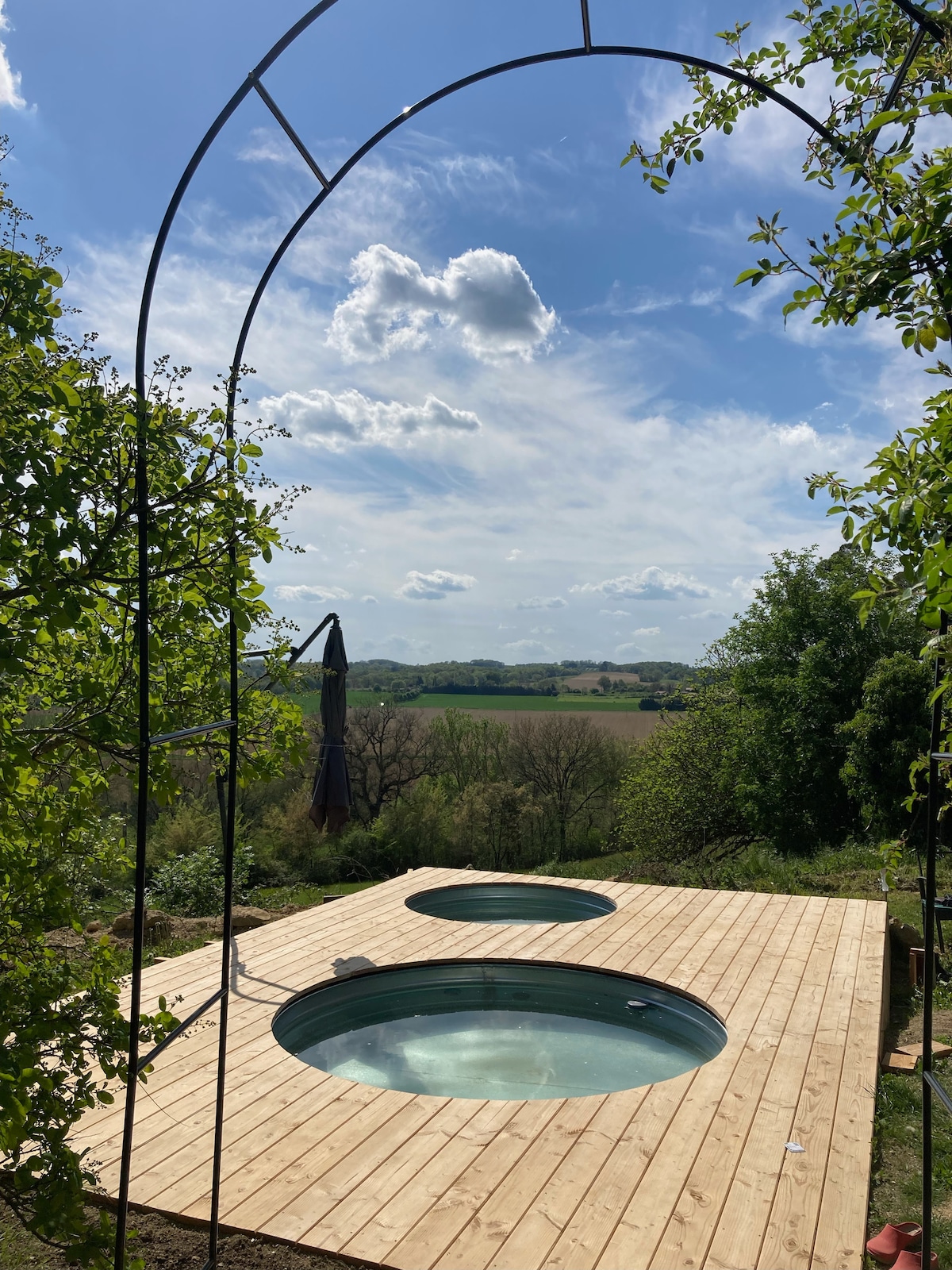 Two circular hot tubs are situated on a wooden deck, surrounded by greenery and open fields. An archway frames the scene, offering a view of the clouds and the serene landscape beyond. Relaxing outdoor space invites guests to enjoy nature.
