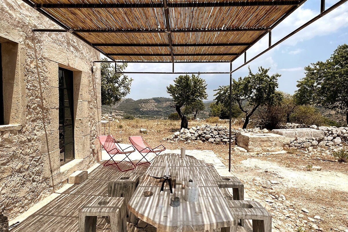 An outdoor dining area features a large wooden table surrounded by rustic chairs, positioned under a shaded pergola. The landscape includes sparse trees and rocky terrain, with distant mountains visible against a clear sky.