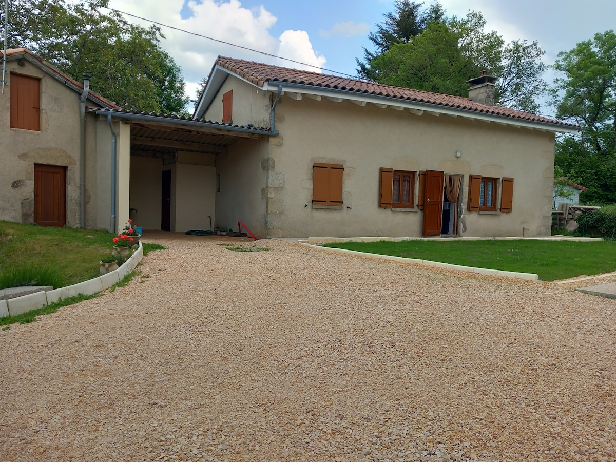 A charming countryside home is showcased with a gravel driveway leading up to the entrance. Soft green grass borders the pathway, and wooden shutters adorn the windows. A covered porch area offers shelter, with trees providing a natural backdrop.