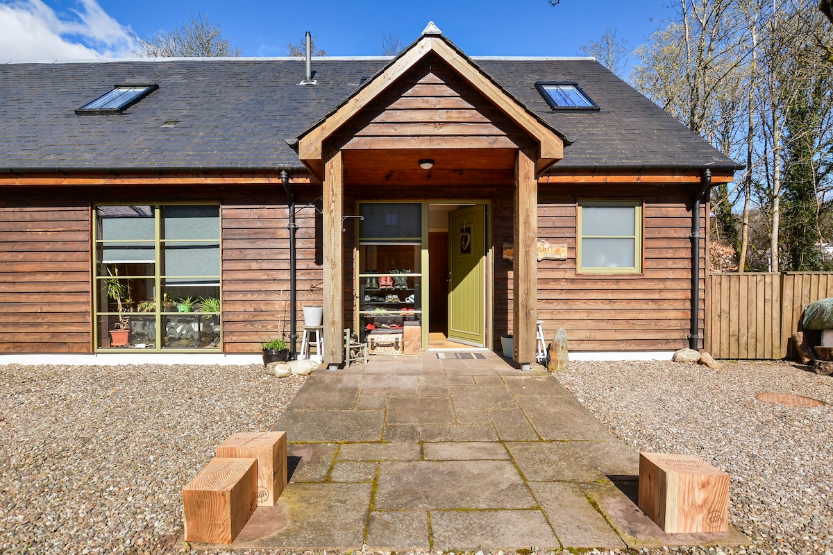 The entrance to the house features a stone pathway leading to a welcoming porch, flanked by large windows. A wooden door with glass inserts provides access to the interior. Two wooden seating blocks are positioned on the ground, complemented by a gravel area.