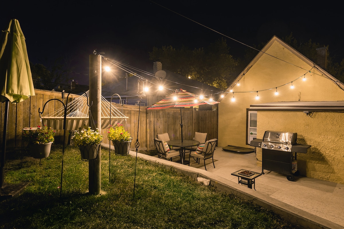 A private backyard patio features subtle lighting from string lights overhead, illuminating a seating area with a table and chairs. A hammock and a grill are also present, surrounded by hanging flower planters and well-maintained grass.