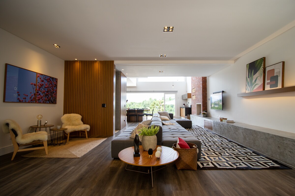 A spacious living area is illuminated by natural light flowing through large windows. A contemporary sectional sofa is positioned alongside a round coffee table adorned with decorative items. Complementary art pieces adorn the walls, while a cozy armchair adds to the inviting atmosphere.