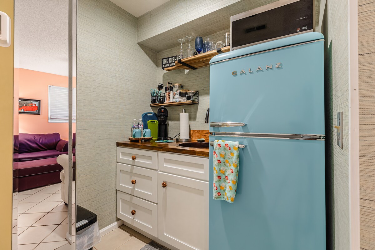 A compact kitchen area is equipped with a retro blue refrigerator and white cabinetry. Light-colored countertops hold kitchen essentials, while a coffee maker and glassware are arranged on the shelf above. Soft overhead lighting highlights the space, creating an organized and functional environment.