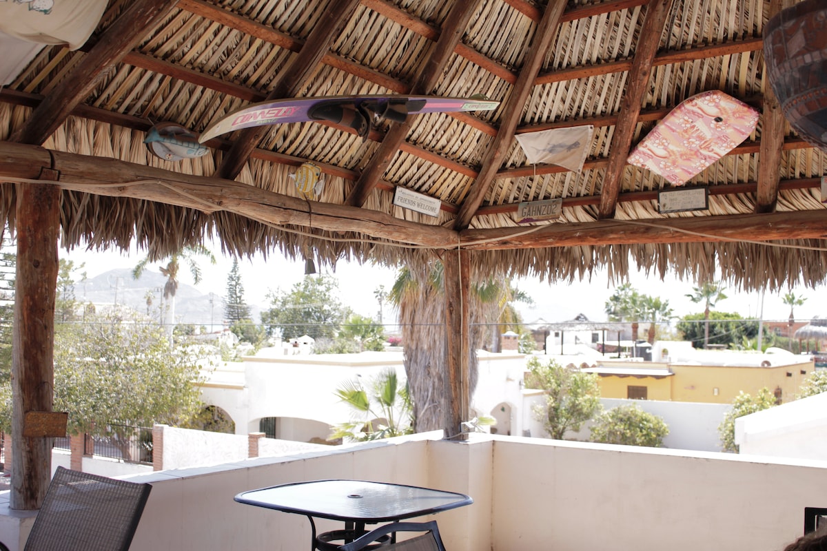 A private terrace is covered with traditional thatch, featuring wooden beams and surfboards mounted decoratively on the walls. A round table and chairs are positioned for outdoor seating, surrounded by views of nearby palm trees and rooftops of local buildings under a clear sky.