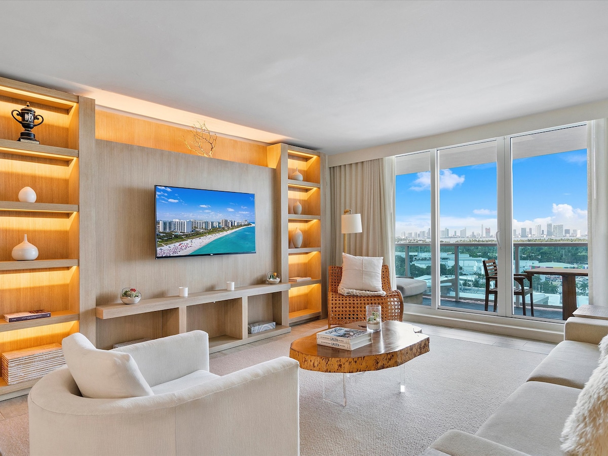 A spacious living room features a large Smart TV mounted on a wooden entertainment unit with illuminated shelves. A comfortable white sofa and a woven chair are arranged around a unique coffee table, while expansive windows offer views of the city skyline and the Intracoastal.