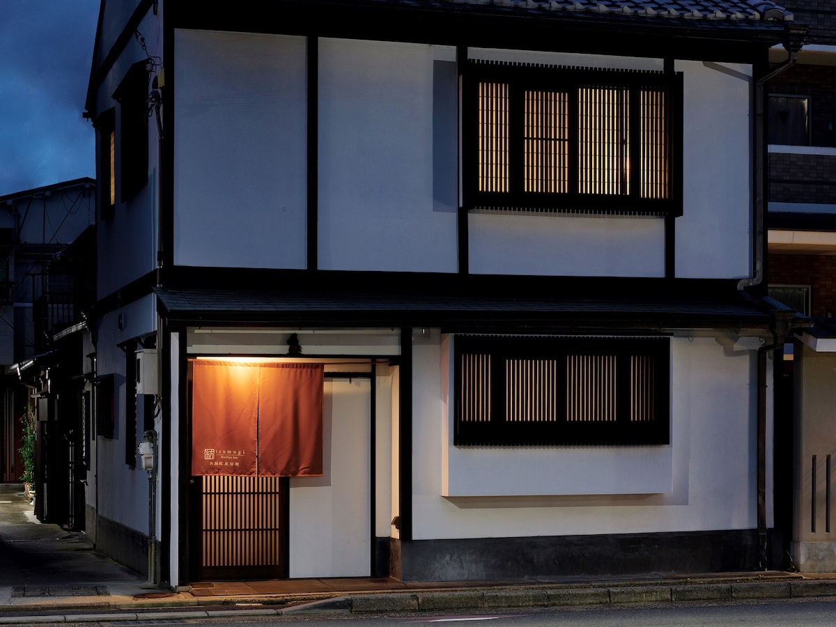 The exterior of a traditional Kyoto townhouse is shown, featuring white walls and dark wooden accents. Vertical wooden slats cover the windows, allowing glimpses inside. A soft orange light emanates from the entrance, creating a welcoming contrast against the night sky.